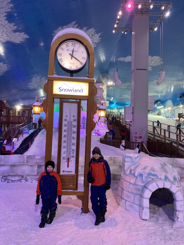 duas crianças ao lado de um grande termômetro e espaço de neve artificial ao fundo