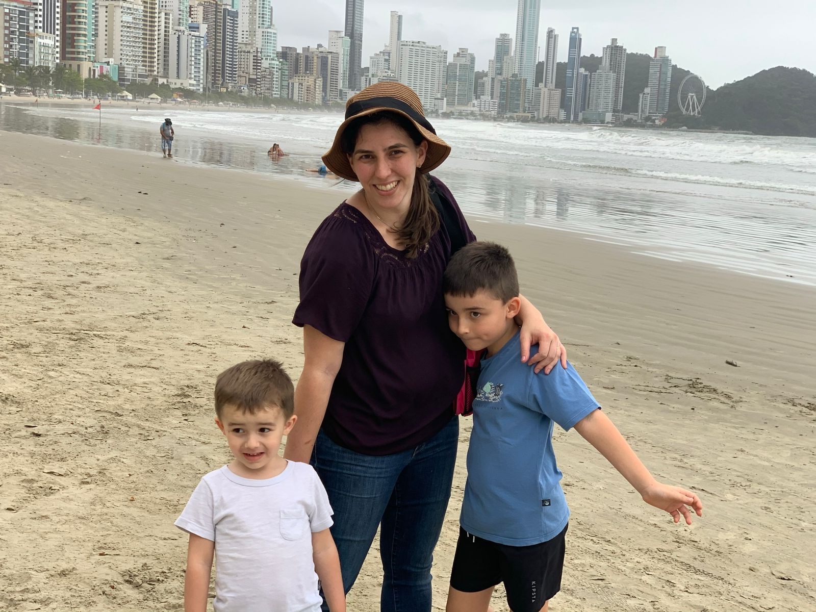mãe e dois filhos juntos a beira da praia com prédios ao fundo de balneário camboriu