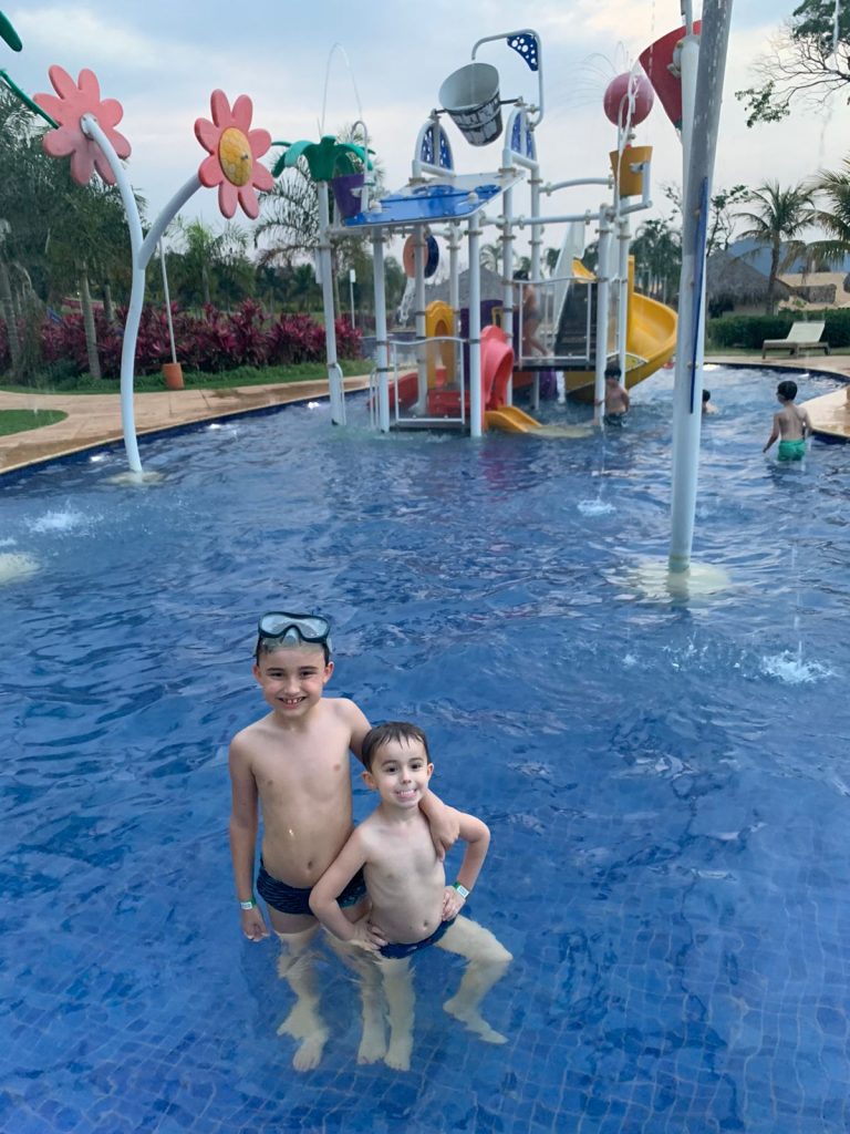 dois meninos pequenos dentro da piscina abraçados com parque aquático infantil no fundo
