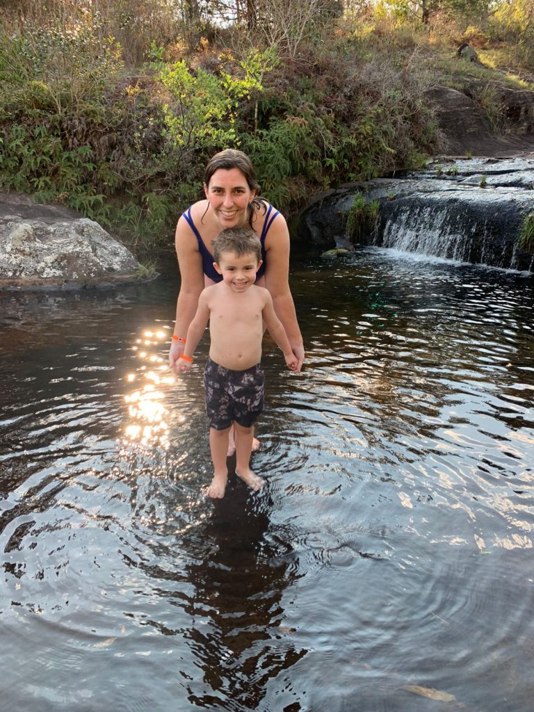 criança ne frente da mãe em um rio raso com cachoeira pequena ao fundo