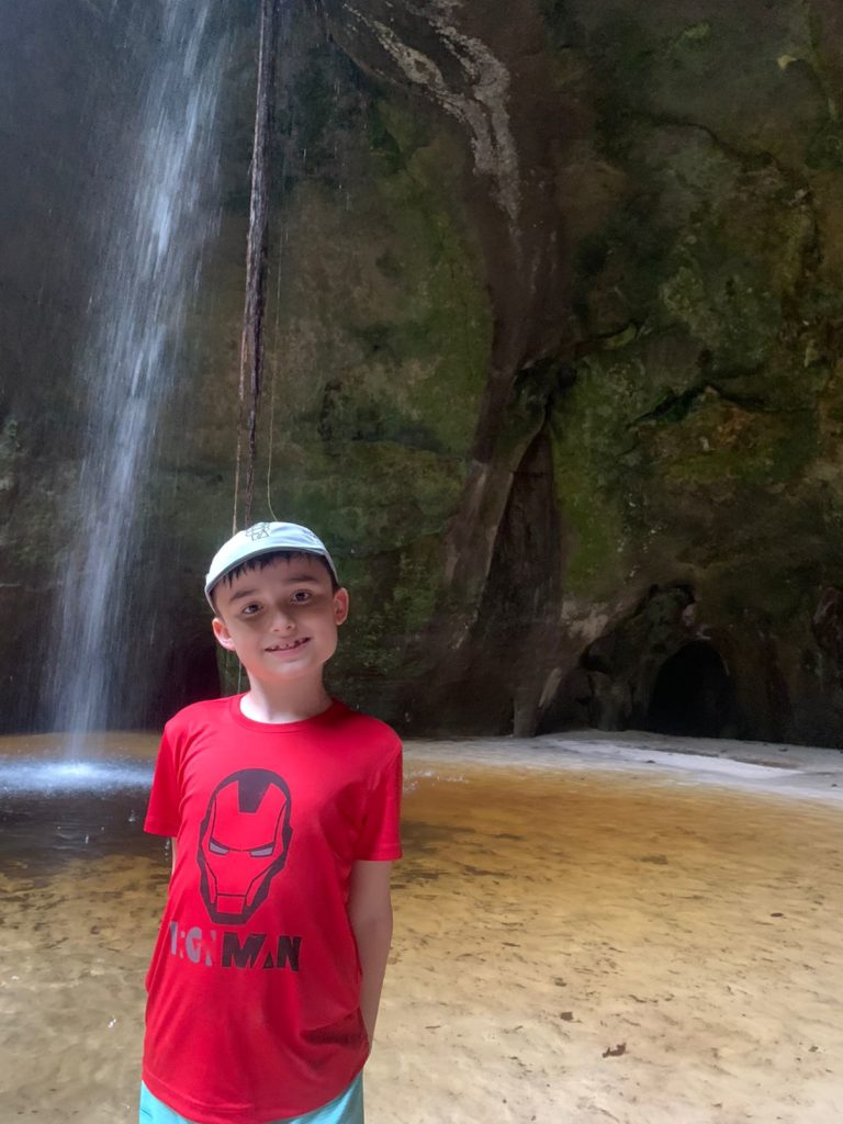 menino sorrindo em frente a pequena cachoeira com gruta no fundo