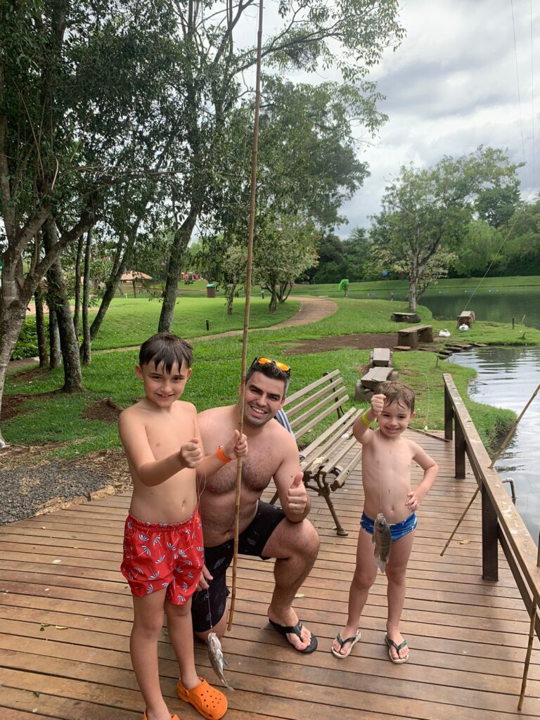 pai e dois filhos com vara de pesca na mão ao lado de um lago