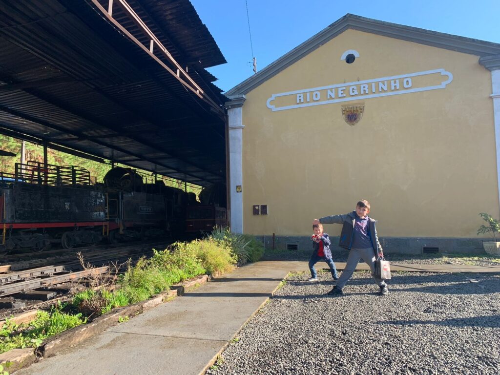 dois meninos em frente a fachada lateral de estação de trem com letreiro rio negrinho