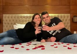 Homem e mulher deitados de lado em cama com pétalas de rosas fazendo um brinde