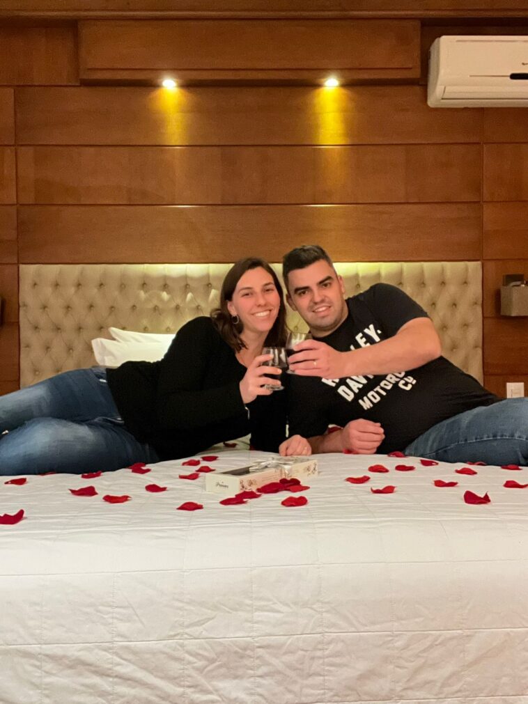 Homem e mulher deitados de lado em cama com pétalas de rosas fazendo um brinde