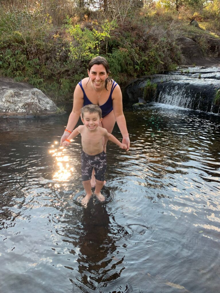 mãe e filho pequeno em cima de parte rasa de rio com pequena cachoeira ao fundo.