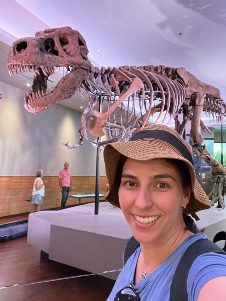 selfie de mulher sorrindo com esqueleto de t-rex ao fundo