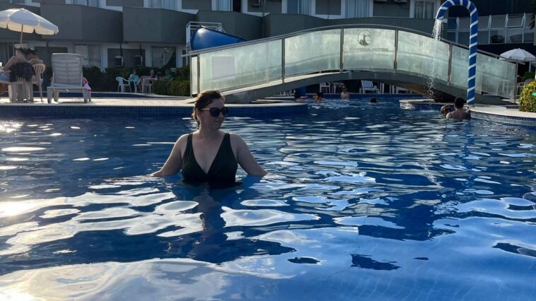 mulher dentro de grande piscina com ponte e prédio de hotel ao fundo