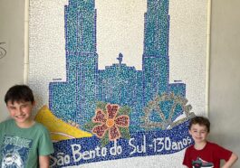dois meninos em frente a mural de mosaico de igreja e título são bento do sul 130 anos