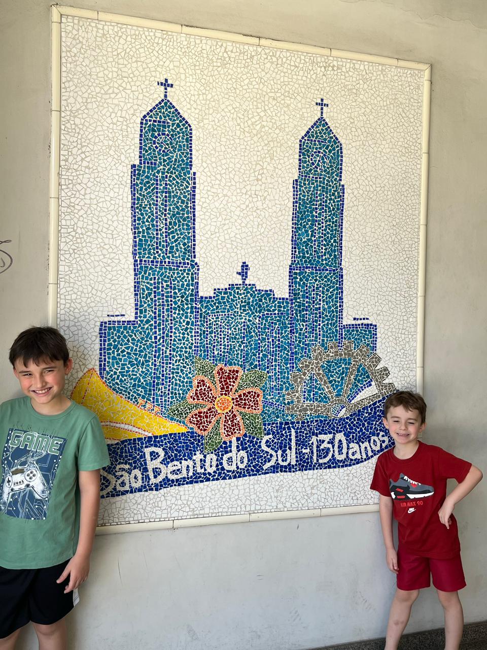 dois meninos em frente a mural de mosaico de igreja e título são bento do sul 130 anos