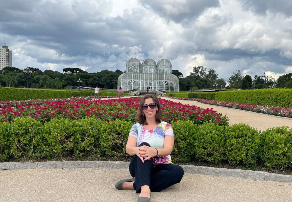 mulher sentada em frente a jardim com estufa do jardim botânico de Curitiba ao fundo.