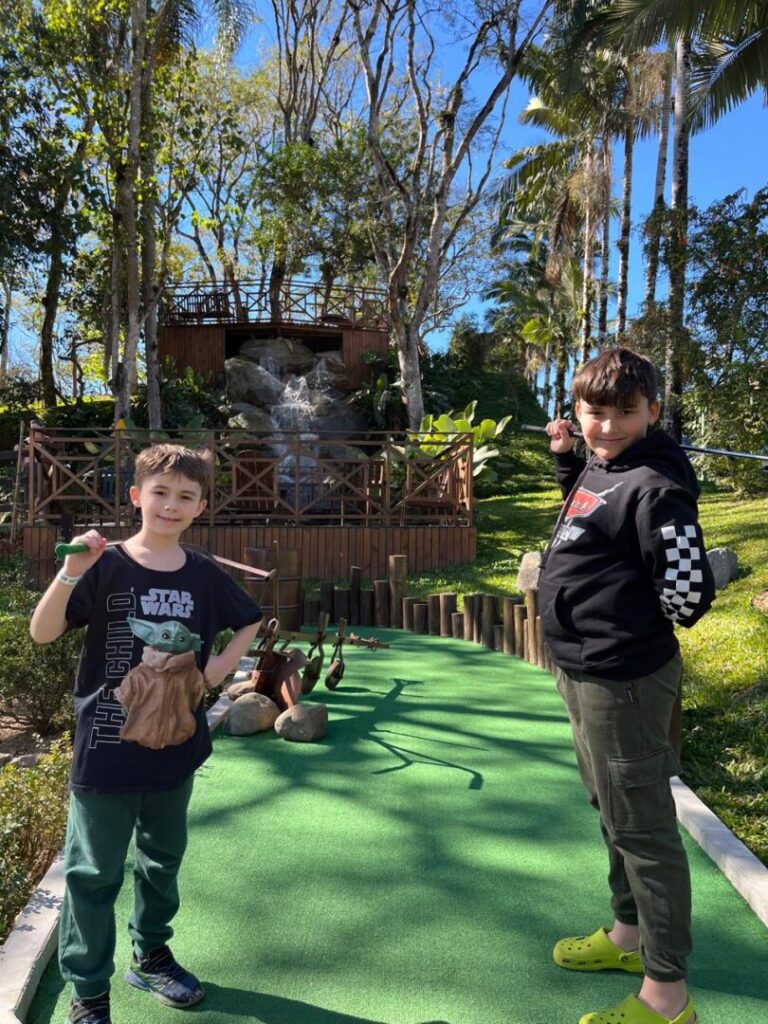 Dois meninos em pista de mini golfe com cachoeira ao fundo.